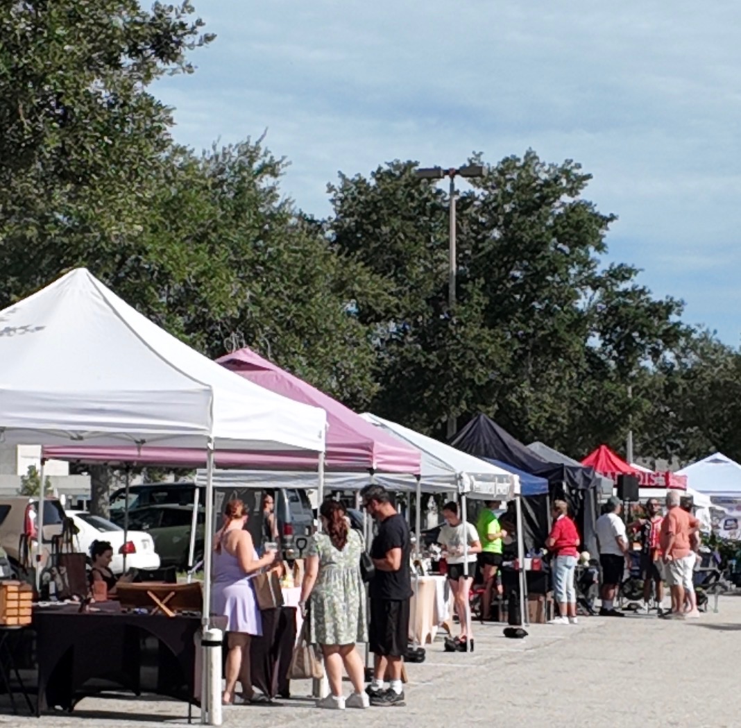A long row of white and pink market tents with shoppers browsing tables under the trees on a sunny Saturday morning